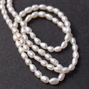 May include: A strand of white, oval-shaped freshwater pearls strung together. The pearls exhibit a natural, lustrous finish with slight variations in their individual shapes and sizes, creating a continuous, flowing line of white beads.