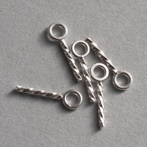 May include: A collection of four silver-toned metal jewelry findings, each featuring a circular loop attached to a slender, twisted bar. These small components have a polished, reflective finish and are arranged in a scattered, overlapping pattern.