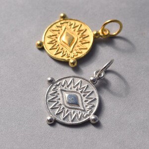 May include: Two gold and silver pendants with a stylized eye design. The pendants are round and have a textured surface. The eye design is surrounded by a starburst pattern.