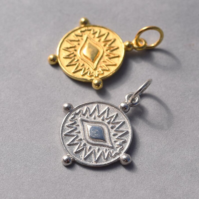 May include: Two gold and silver pendants with a stylized eye design. The pendants are round and have a textured surface. The eye design is surrounded by a starburst pattern.