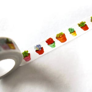 Peut inclure: Un rouleau de ruban adhésif washi blanc avec un motif répété de succulentes en pot de différentes couleurs. Le ruban est partiellement déroulé, révélant le motif.