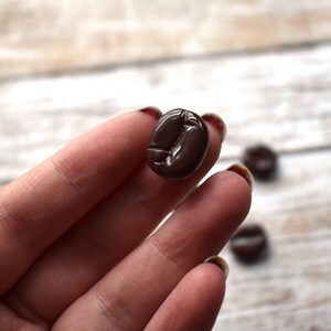 May include: A close-up of a brown coffee bean charm. The charm is being held by a hand with manicured nails.