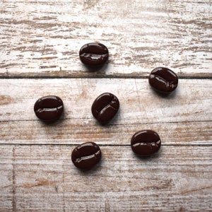 May include: Six brown coffee beans on a white wooden surface.