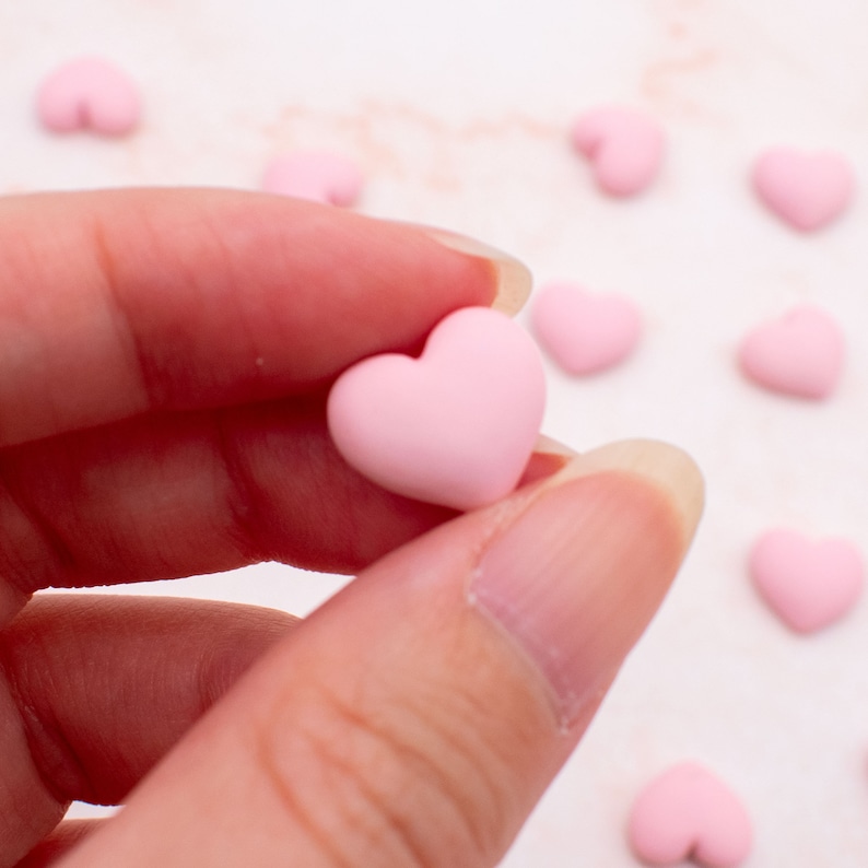 K&ouml;nnte beinhalten: Nahaufnahme eines rosa herzf&ouml;rmigen Charms, der in einer Hand gehalten wird. Der Charm ist klein und glatt und von anderen rosa herzf&ouml;rmigen Charms umgeben.