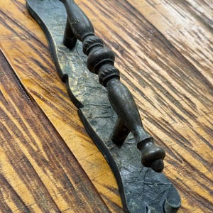May include: Antique bronze door handle with an ornate design. The handle has a detailed, textured surface and a classic, vintage aesthetic. The handle is mounted on a wooden surface.