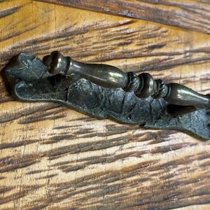 May include: A bronze-toned drawer pull with a decorative leaf-shaped backplate. The handle has a detailed, ornate design. The pull is mounted on a wooden surface with visible grain.