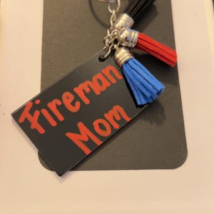 May include: A keychain featuring a black tag with the words "Fireman Mom" in red. The keychain has a silver ring, black, red, and blue tassels, and silver detailing.