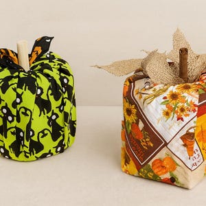 Puede incluir: Dos calabazas de tela para la decoración de otoño. Una es verde con siluetas de gatos negros, un tallo de madera y hojas de tela naranja y negra. La otra es un patchwork de telas con temática otoñal.