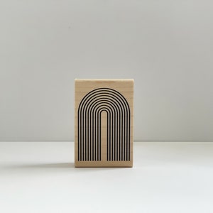 K&ouml;nnte beinhalten: Ein Holzstempel mit einem schwarzen Bogendesign. Das Design zeigt eine Reihe von gebogenen Linien, die ein minimalistisches, geometrisches Muster bilden. Der Stempel ist rechteckig und aus hellem Holz gefertigt.