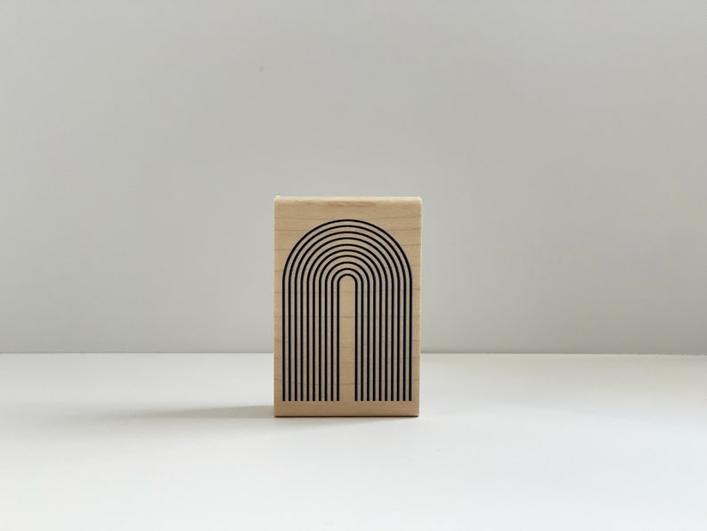 K&ouml;nnte beinhalten: Ein Holzstempel mit einem schwarzen Bogendesign. Das Design zeigt eine Reihe von gebogenen Linien, die ein minimalistisches, geometrisches Muster bilden. Der Stempel ist rechteckig und aus hellem Holz gefertigt.