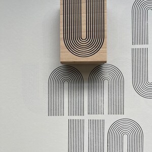 K&ouml;nnte beinhalten: Ein Holzstempel mit einem schwarzen, bogenf&ouml;rmigen Design in Form des Buchstabens "U". Der Stempel ist auf eine wei&szlig;e Oberfl&auml;che gedr&uuml;ckt, wodurch mehrere Abdr&uuml;cke desselben Designs entstehen. Das Design ist eine Reihe paralleler Linien.