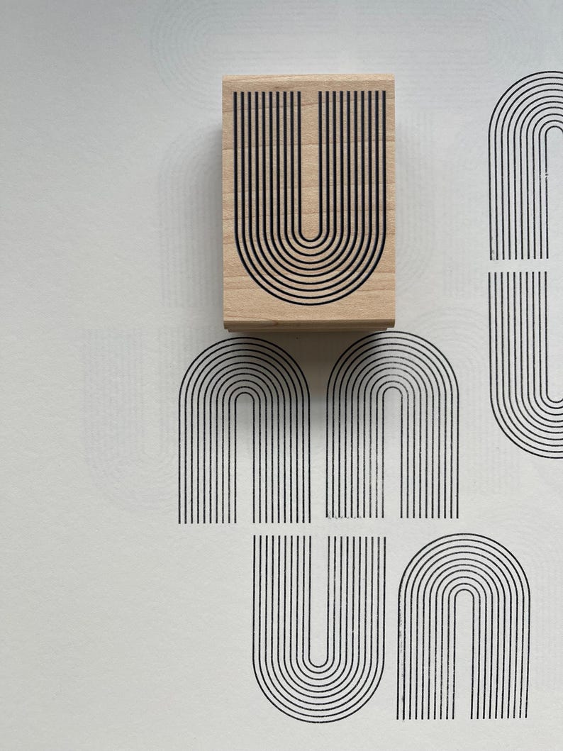 K&ouml;nnte beinhalten: Ein Holzstempel mit einem schwarzen, bogenf&ouml;rmigen Design in Form des Buchstabens "U". Der Stempel ist auf eine wei&szlig;e Oberfl&auml;che gedr&uuml;ckt, wodurch mehrere Abdr&uuml;cke desselben Designs entstehen. Das Design ist eine Reihe paralleler Linien.
