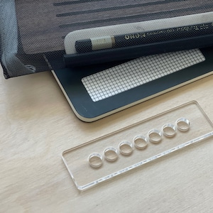 May include: A clear acrylic pencil holder with eight round holes for storing pencils. The holder is on a light wood surface.