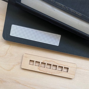 May include: A wooden ruler with nine evenly spaced rectangular cutouts. The ruler is light brown and has a natural wood grain.