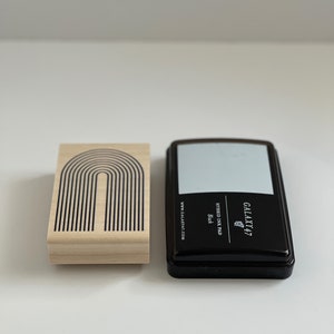 K&ouml;nnte beinhalten: Ein Holzstempel mit einem schwarzen Bogendesign neben einem schwarzen Stempelkissen. Das Stempelkissen tr&auml;gt den Text "GALAXY 47" und "HYBRID INK PAD Black". Der Stempel hat die Website "GALAXY47.COM" aufgedruckt.