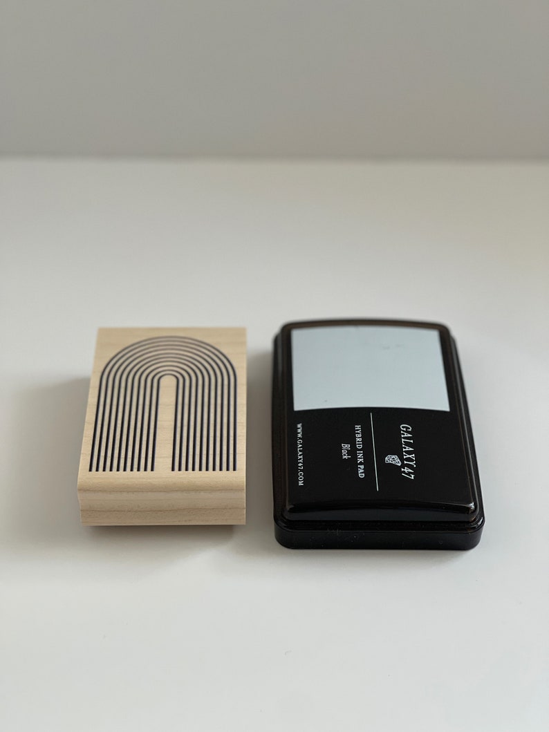 K&ouml;nnte beinhalten: Ein Holzstempel mit einem schwarzen Bogendesign neben einem schwarzen Stempelkissen. Das Stempelkissen tr&auml;gt den Text "GALAXY 47" und "HYBRID INK PAD Black". Der Stempel hat die Website "GALAXY47.COM" aufgedruckt.