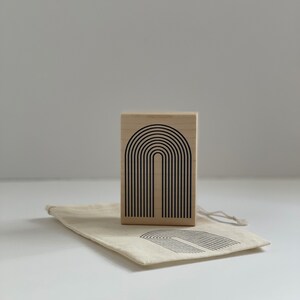 K&ouml;nnte beinhalten: Ein Holzstempel mit einem schwarzen Bogendesign, begleitet von einem passenden Canvas-Beutel. Der Stempel zeigt eine Reihe von gebogenen Linien, die einen Bogen bilden. Der Beutel hat ein &auml;hnliches Design aufgedruckt.