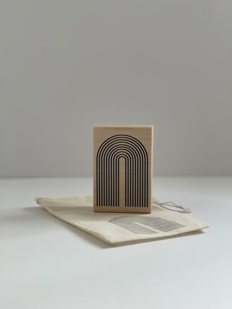 K&ouml;nnte beinhalten: Ein Holzstempel mit einem schwarzen Bogendesign, begleitet von einem passenden Canvas-Beutel. Der Stempel zeigt eine Reihe von gebogenen Linien, die einen Bogen bilden. Der Beutel hat ein &auml;hnliches Design aufgedruckt.