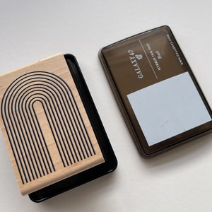 K&ouml;nnte beinhalten: Ein Holzstempel mit einem schwarzen Bogendesign und einem schwarzen Stempelkissen. Das Stempelkissen ist mit "GALAXY 47 HYBRID INK PAD Black" beschriftet. Der Stempel hat eine helle Holzmaserung.