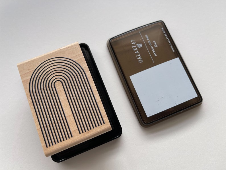 K&ouml;nnte beinhalten: Ein Holzstempel mit einem schwarzen Bogendesign und einem schwarzen Stempelkissen. Das Stempelkissen ist mit "GALAXY 47 HYBRID INK PAD Black" beschriftet. Der Stempel hat eine helle Holzmaserung.