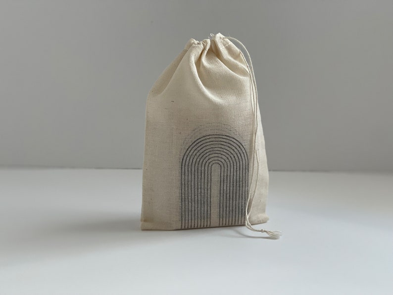 K&ouml;nnte beinhalten: Ein nat&uuml;rlicher Canvas-Kordelzugbeutel mit einem schwarzen Bogendesign. Der Beutel wird mit einem Kordelzug verschlossen und steht auf einer wei&szlig;en Oberfl&auml;che. Der Beutel besteht aus einem strukturierten Stoff.