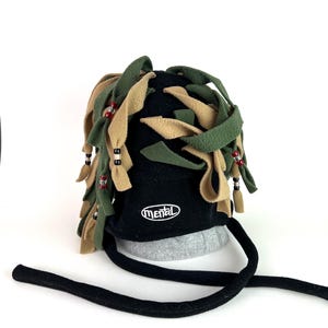 Puede incluir: Un sombrero negro con un diseño único, con tiras de tela verdes y beige y detalles de cuentas rojas. La palabra "mental" está bordada en la parte delantera. Una correa negra se extiende desde el sombrero.