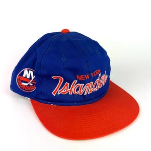 Könnte beinhalten: Blau-rote Baseballkappe mit dem Schriftzug "New York Islanders" in weiß. Die Kappe hat einen roten Schirm und einen kleinen orangefarbenen Knopf oben. Das Teamlogo befindet sich an der Seite.