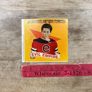 May include: Square sticker featuring a young person in a red and black top with the text "EVIL EMPIRE" on a banner. The sticker has a yellow background with a red star and the text "RAGE AGAINST THE MACHINE".