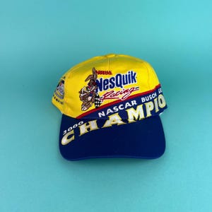 Puede incluir: Gorra de béisbol amarilla y azul marino con el logotipo de Nesquik y el conejo de Nesquik sosteniendo una bandera a cuadros. La gorra también tiene el texto "NASCAR BUSCH SERIES CHAMPION 2000".