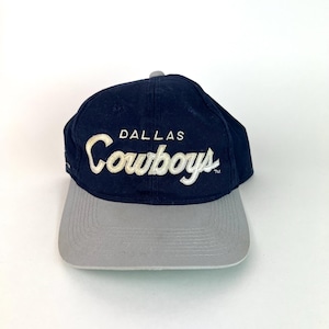 May include: Navy blue and gray baseball cap with the word "Dallas Cowboys" embroidered in white script. The cap has a curved brim and a gray adjustable strap.