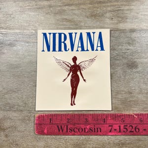 May include: A rectangular sticker with the word "NIRVANA" in blue block letters above a maroon silhouette of a winged figure. A red ruler with white text is at the bottom.