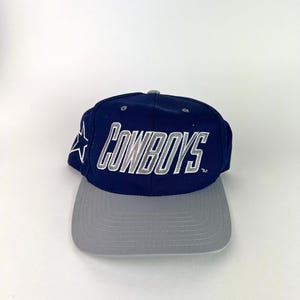 May include: A navy blue and gray baseball cap with the word "COWBOYS" in white and gray lettering. A white star is on the side of the cap. The cap has a gray brim.