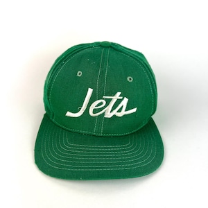 May include: A green baseball cap with a flat brim. The word "Jets" is embroidered in white cursive across the front. The cap is made of a textured fabric and has a classic, structured design.