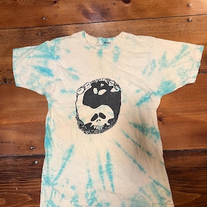 May include: Cream-colored tie-dye t-shirt with a black graphic of a skull and panda bear yin-yang design. The shirt has short sleeves and a crew neck. The tie-dye pattern includes light blue streaks radiating from the center.
