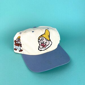 Puede incluir: Gorra de béisbol con corona crema y visera azul. La gorra presenta imágenes bordadas de un personaje de dibujos animados en amarillo, rojo y blanco. Estilo vintage.