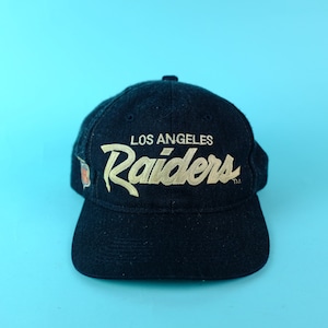 Op de afbeelding: Een zwarte baseballcap met de tekst "LOS ANGELES Raiders" in gouden borduursel. De cap heeft een gebogen klep en een teamlogo aan de zijkant. De achtergrond is lichtblauw.