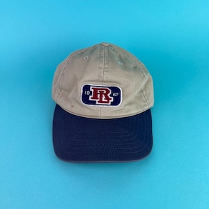 Puede incluir: Gorra de béisbol beige con visera azul marino. La gorra presenta un parche con una "R" roja dentro de un rectángulo azul marino, junto con los números "19" y "67". Fondo azul claro.