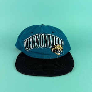 May include: A teal and black baseball cap featuring the word "JACKSONVILLE" in white and black lettering. The cap also has a yellow and black jaguar logo on the side. The hat has a black brim and a teal crown.