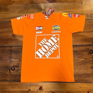 Op de afbeelding: Oranje t-shirt met sponsorlogo's en het Home Depot-logo. Het shirt heeft een ronde hals en korte mouwen. Logo's zijn onder meer Champion, ARP en Nextel Cup Series.