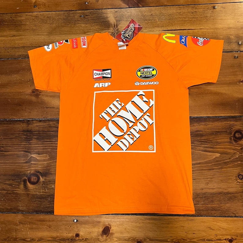 Op de afbeelding: Oranje t-shirt met sponsorlogo's en het Home Depot-logo. Het shirt heeft een ronde hals en korte mouwen. Logo's zijn onder meer Champion, ARP en Nextel Cup Series.