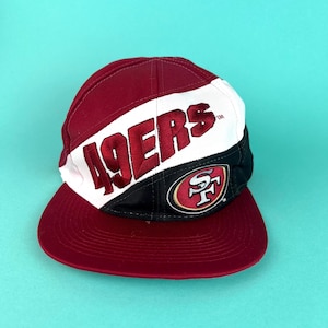 May include: A maroon, white, and black baseball cap with the word "49ERS" embroidered in red. The cap features the team's logo on the side and a maroon brim. The hat is set against a light blue background.