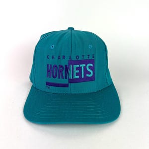 May include: Teal baseball cap with the text "CHARLOTTE HORNETS" embroidered in purple. The cap has a curved bill and a button on top. The hat is a classic style, likely from the 1990s.