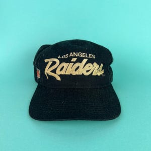 以下が含まれることがあります： 黒い野球帽。前面に金色の文字で「Los Angeles Raiders」と書かれています。つばはカーブしており、クラウンは構造化されています。レイダースのロゴが前面に表示されています。
