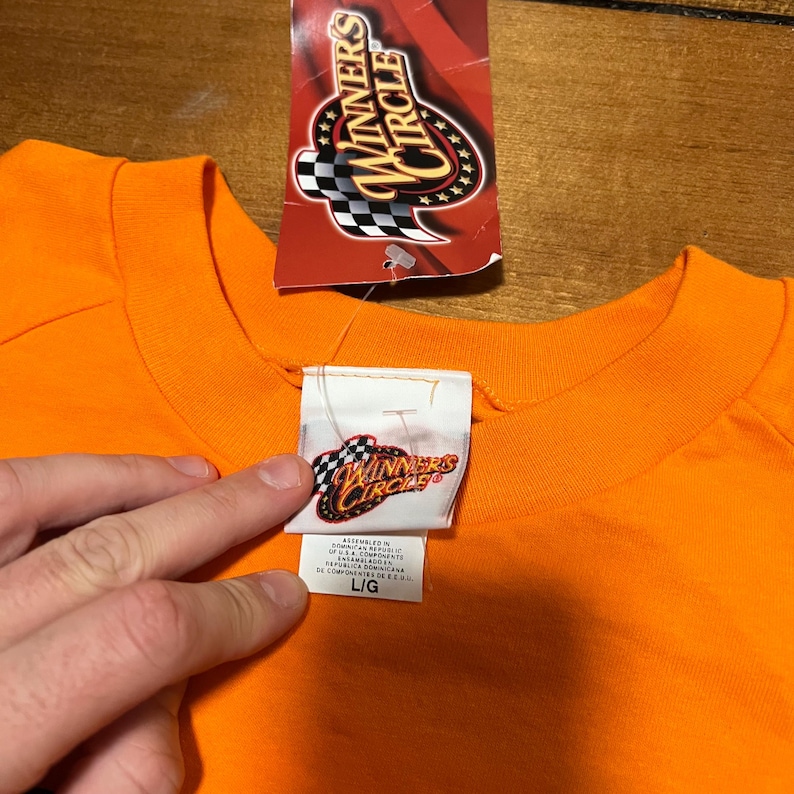 Op de afbeelding: Fel oranje crewneck T-shirt met een "Winners Circle" label eraan. Het label is rood met een geblokt vlagontwerp en de woorden "Winners Circle". Het shirtlabel heeft het "Winners Circle" logo en maat L/G.