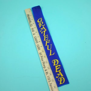 May include: A blue embroidered patch with the words "Grateful Dead" in yellow script. The patch is long and narrow, with a small loop at the top. A wooden ruler is next to the patch.