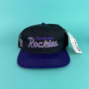 Op de afbeelding: Een zwarte en paarse baseballcap met de tekst "COLORADO Rockies" geborduurd in paars en wit. De cap heeft een paarse klep en een aangehecht label. Sportaccessoire.