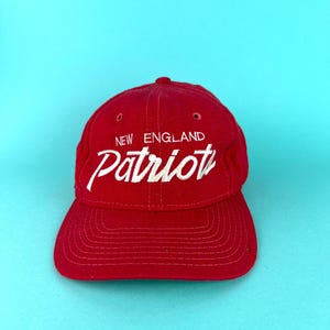 May include: A red baseball cap with the words "NEW ENGLAND Patriots" embroidered in white. The cap has a curved brim and a rounded crown. The hat is a classic style, suitable for sports fans.