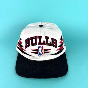 May include: White and black Bulls baseball cap with the team name in red block letters. The cap features the NBA logo and red and black accents. The brim is black.