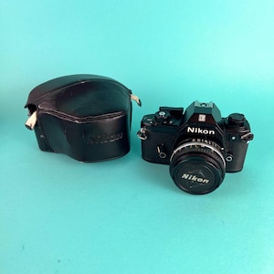 Op de afbeelding: Zwarte Nikon camera met een lensdop en een zwarte leren etui. De camera heeft het Nikon logo op de voorkant en op de lensdop. De etui is ook voorzien van het Nikon logo.