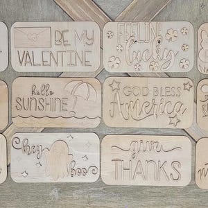 May include: A set of 10 wooden signs with various sayings, including "Happy New Year", "Be My Valentine", "Feeling Lucky", "Honoring All Who Served", "Hello Sunshine", "God Bless America", "It's Fall Y'all", "Cheery Boo", "Give Thanks", and "Merry Christmas".